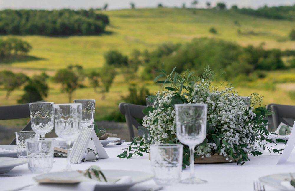 Charming outdoor table setup with flowers and wineglasses against scenic hills backdrop.